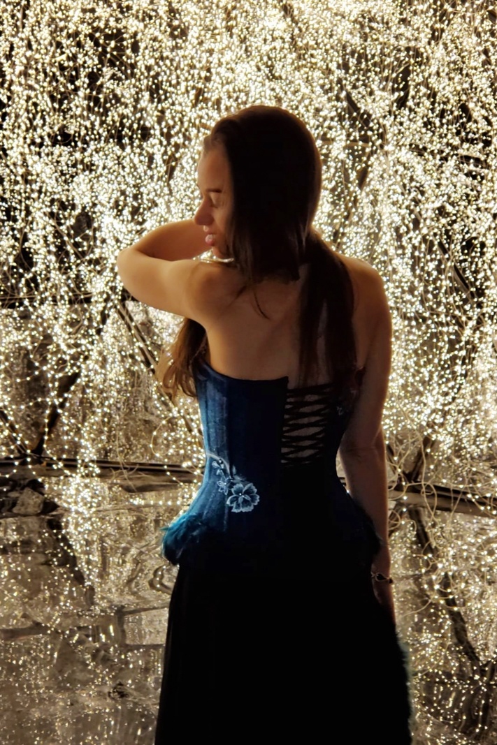 Stella standing in front of a Christmas Tree showing the back of her blue corset.